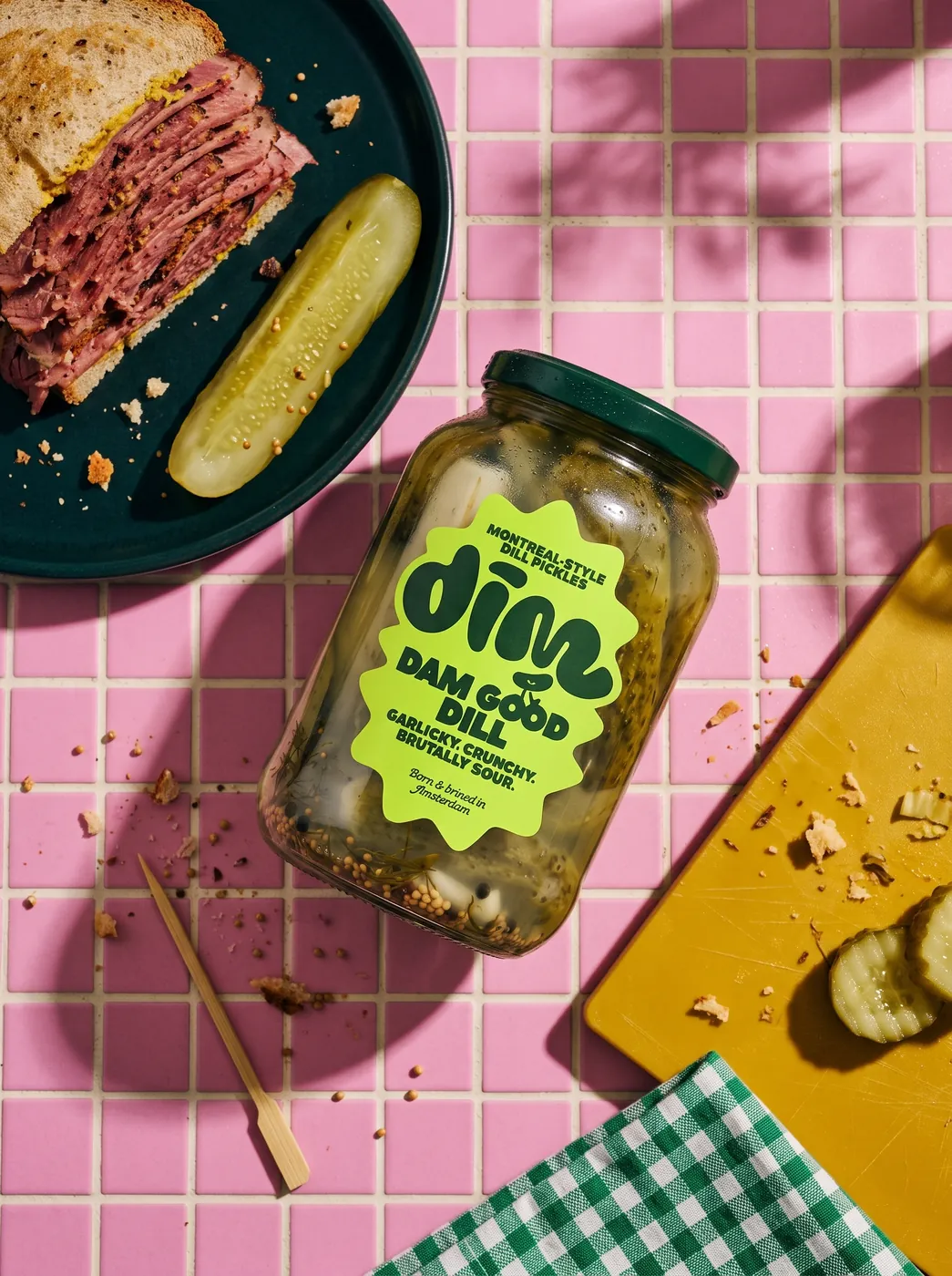 Flatlay of a Dim Pickles jar on pink tiles with a pastrami sandwich and pickle