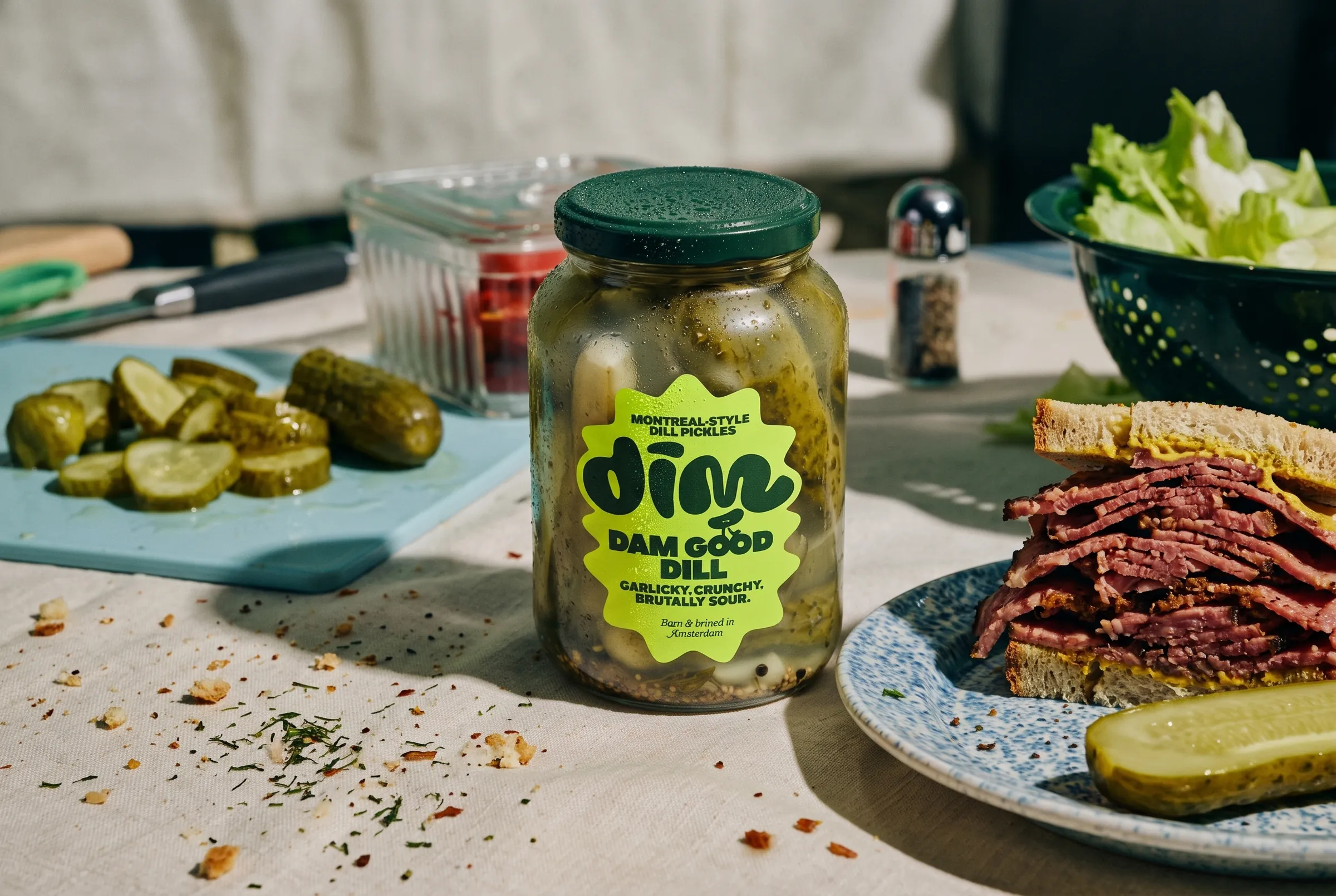 Dim Pickles jar on a linen tablecloth with sliced pickles and a sandwich